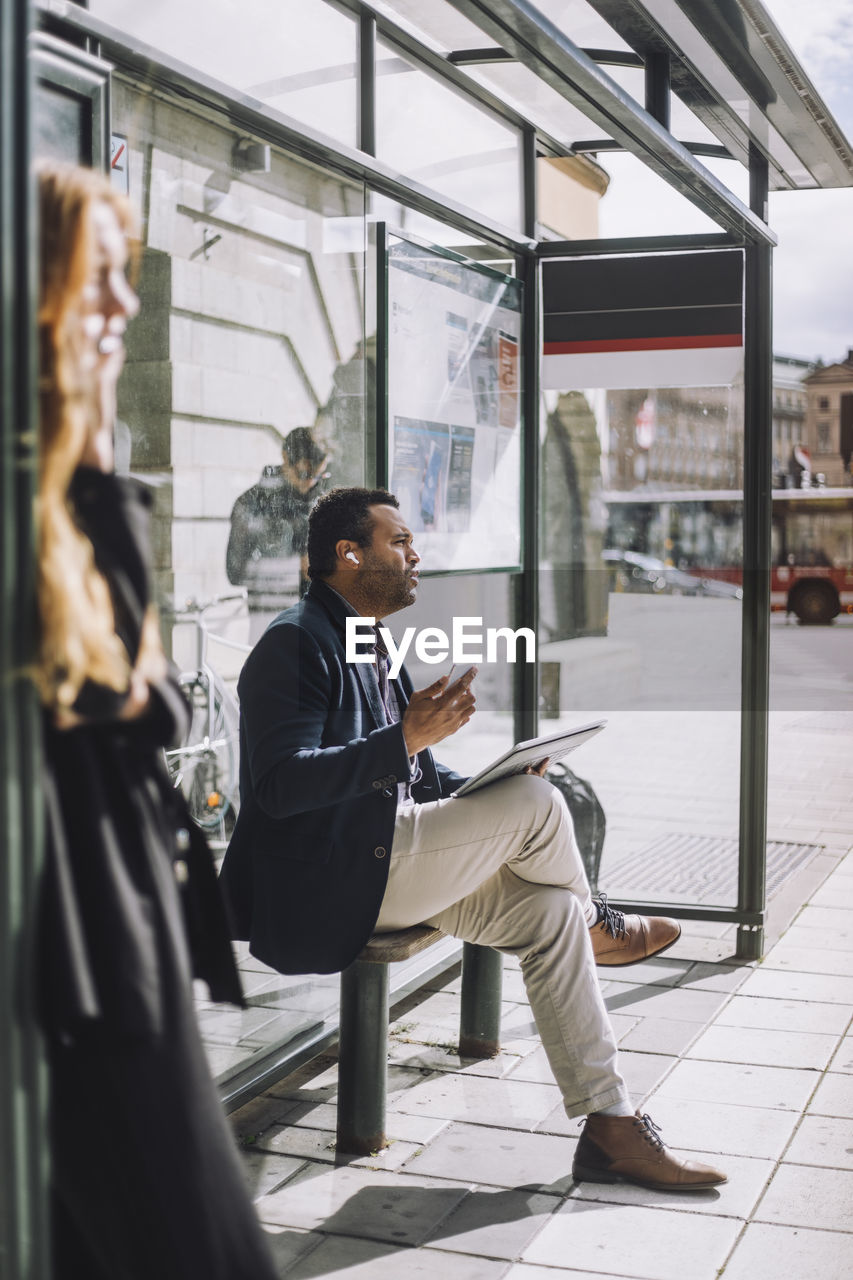 Full length of male professional sitting with laptop at bus stop during sunny day