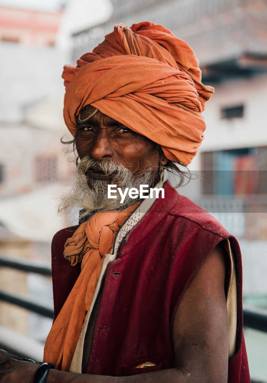 Varanasi, india - february, 2018: aged bearded hindu male in orange turban wearing red sleeveless vest standing on street and looking at camera