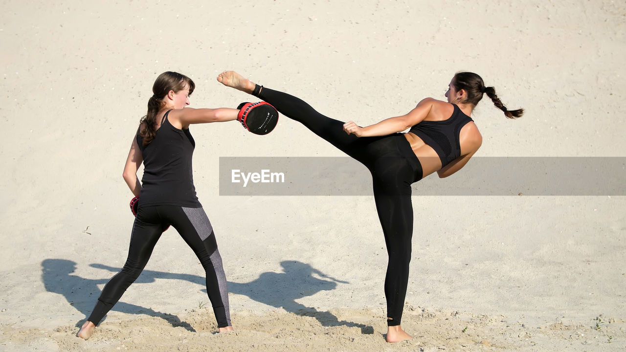 Young women in black fitness suits are engaged in a pair, work out kicks, on a deserted beach