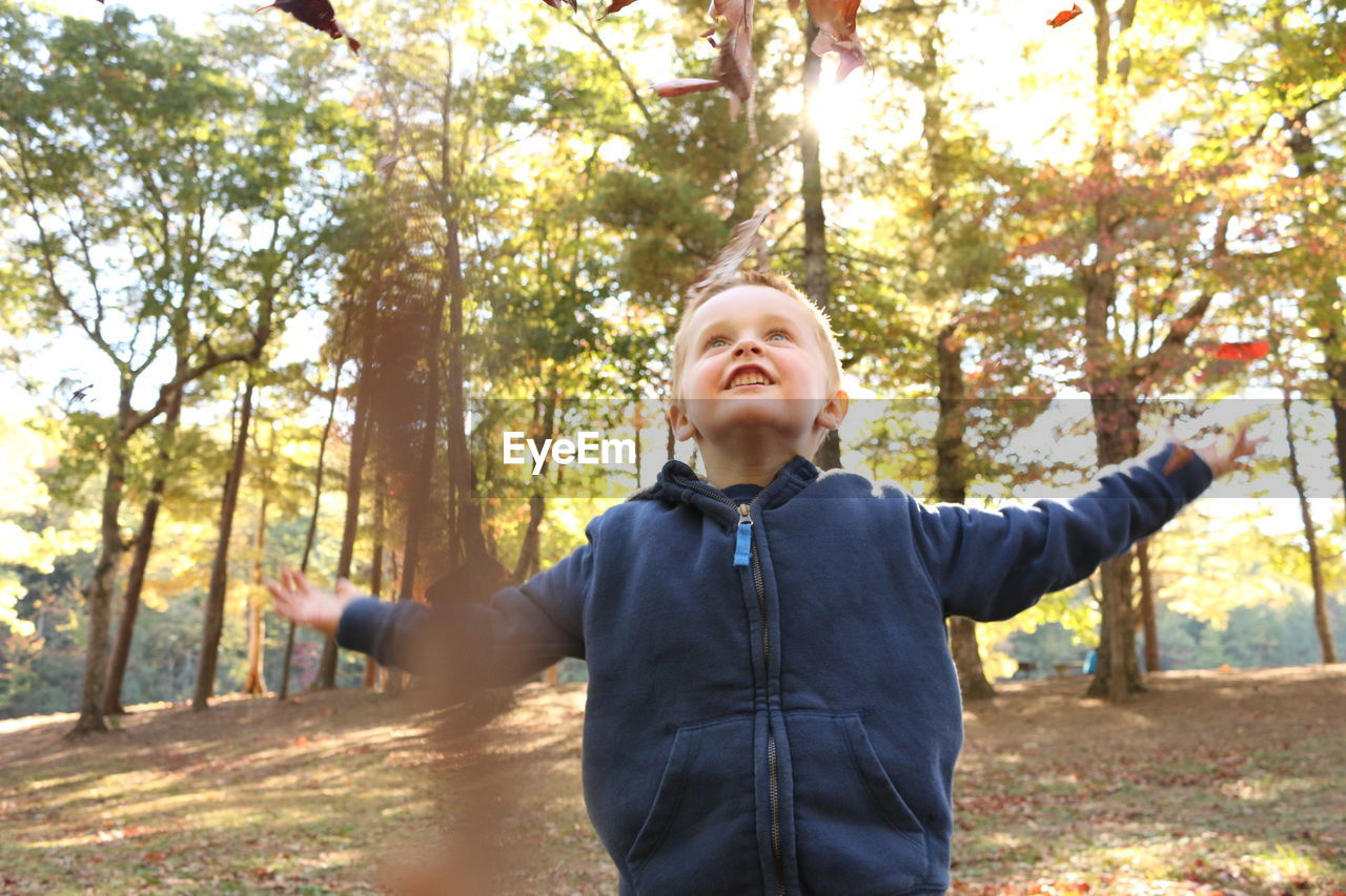 Happy boy throwing leaves while standing in park against tree