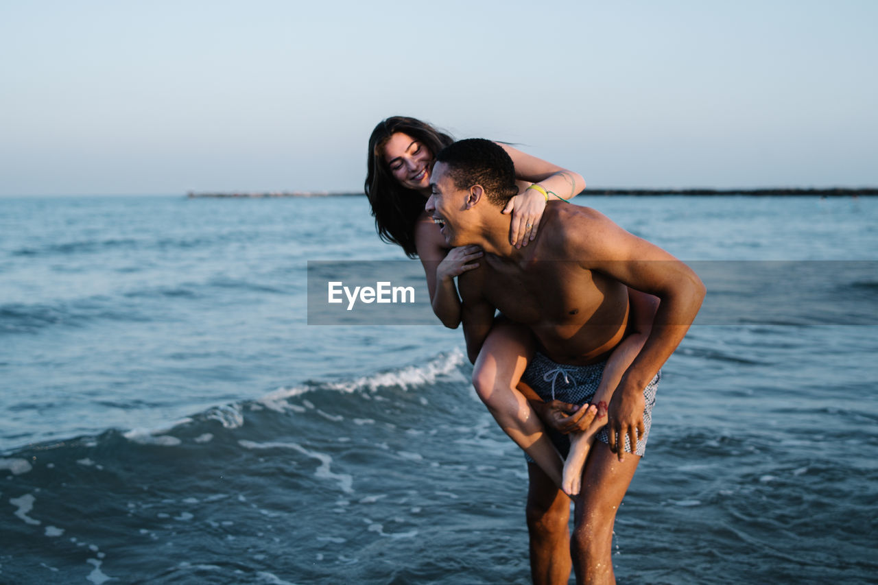 Young happy female tourist riding piggyback on african american boyfriend against wavy ocean during honeymoon