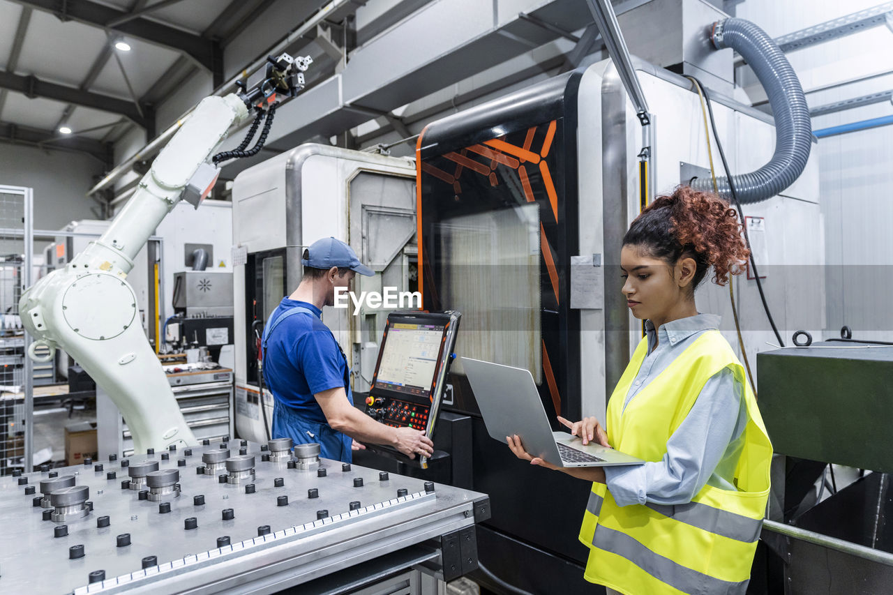 Young engineer using laptop with colleague operating machine in factory