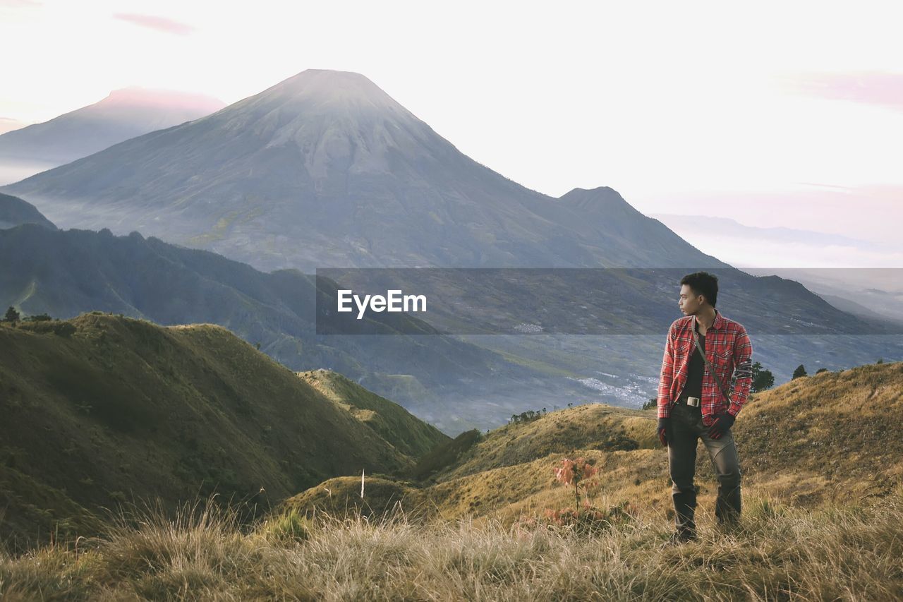 Young man standing on field while looking at mountains against sky