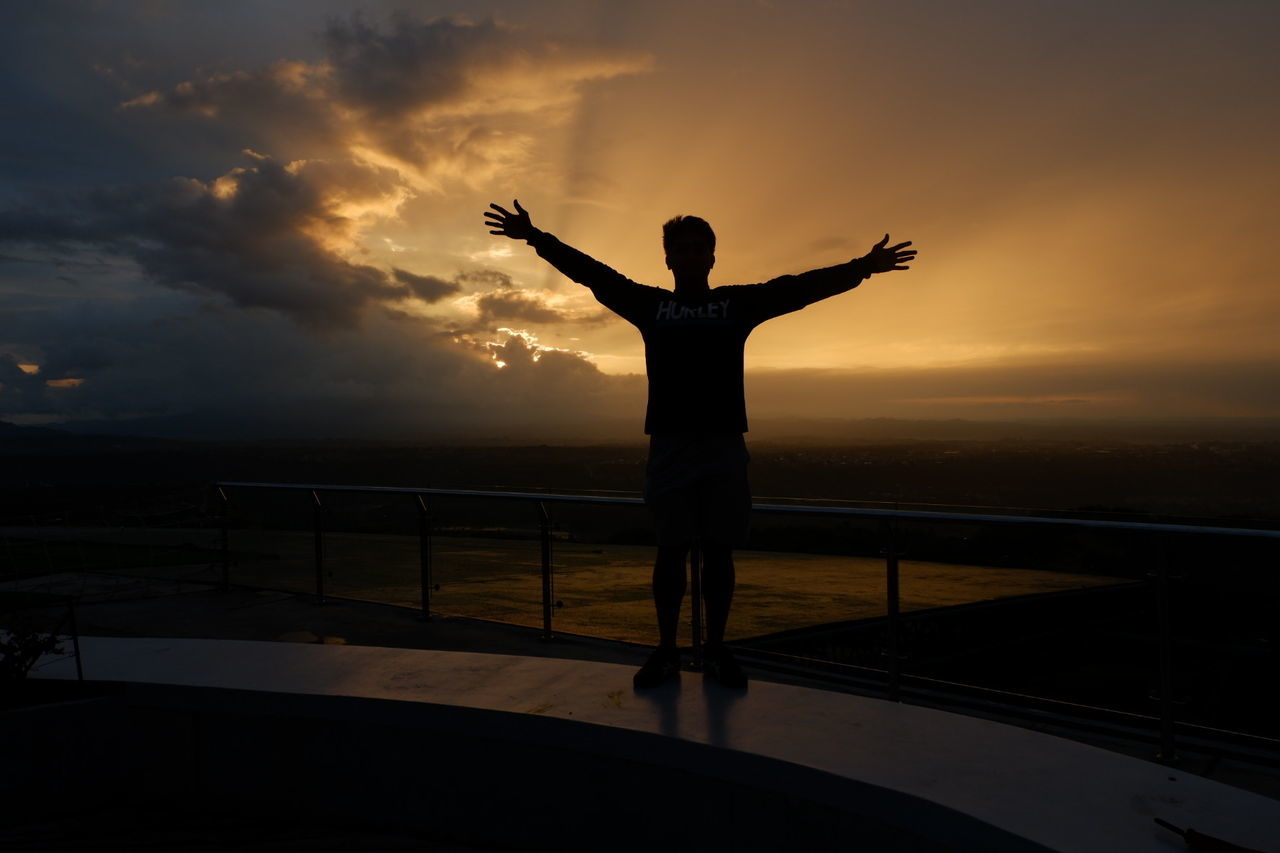 Rear view of silhouette mid adult man with arms outstretched standing against sky during sunset