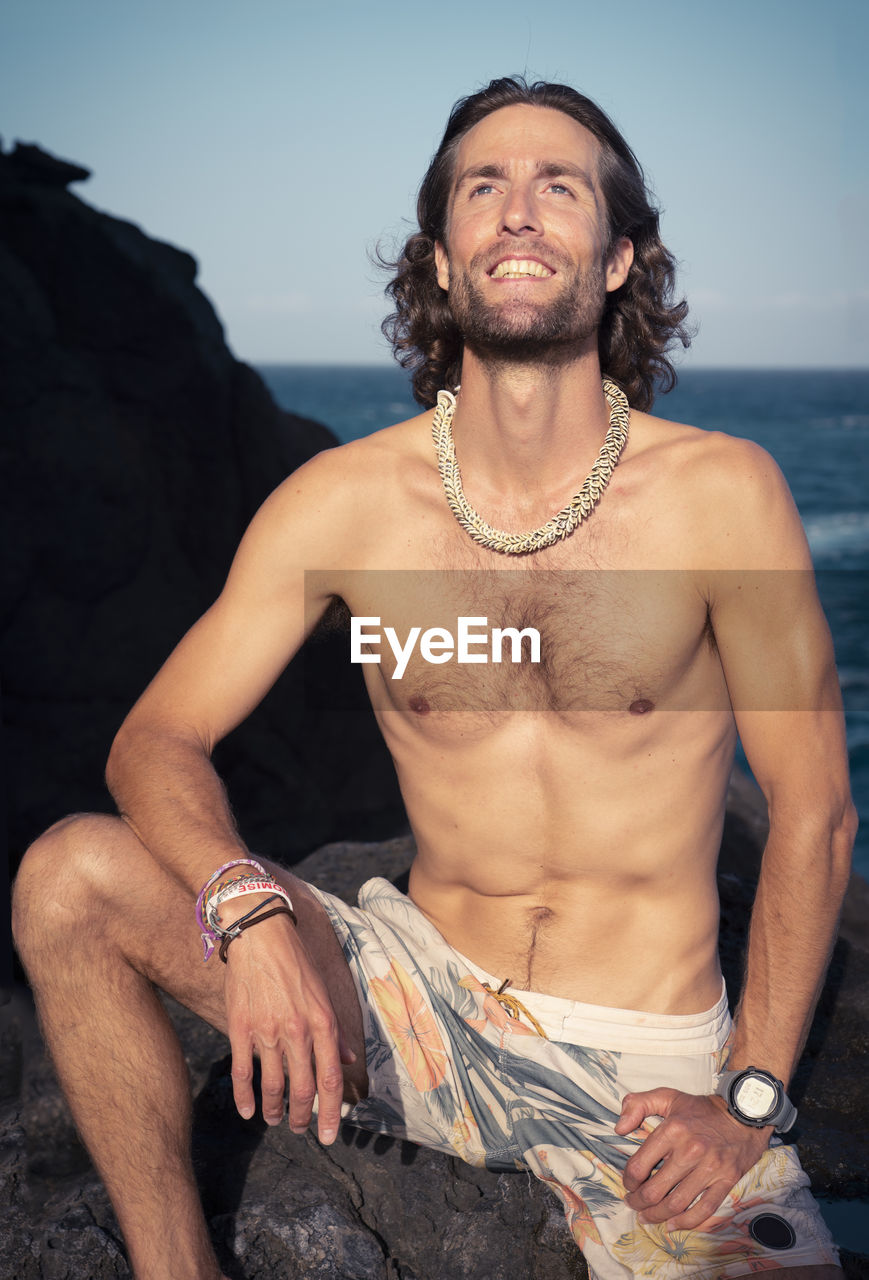 Shirtless man sitting on rock at beach against sky