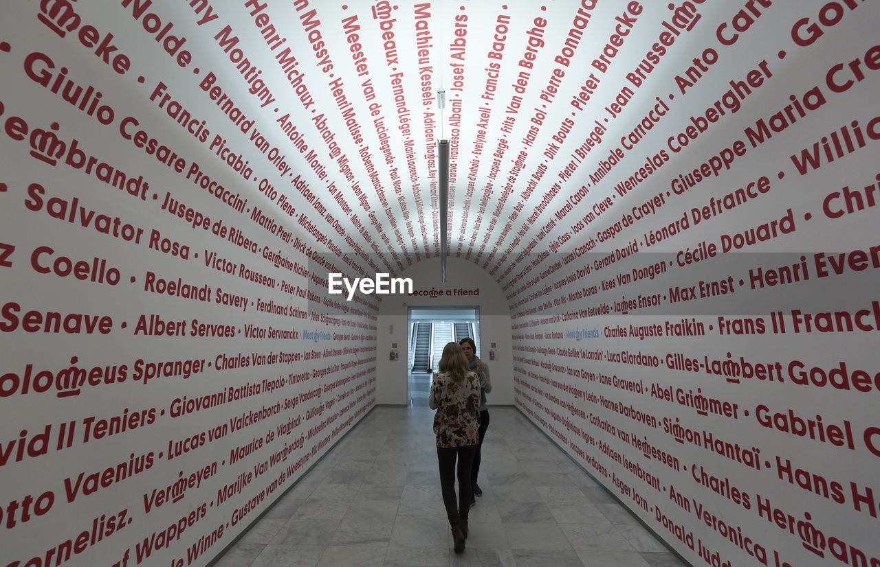 REAR VIEW OF WOMAN WALKING IN CORRIDOR OF PEOPLE