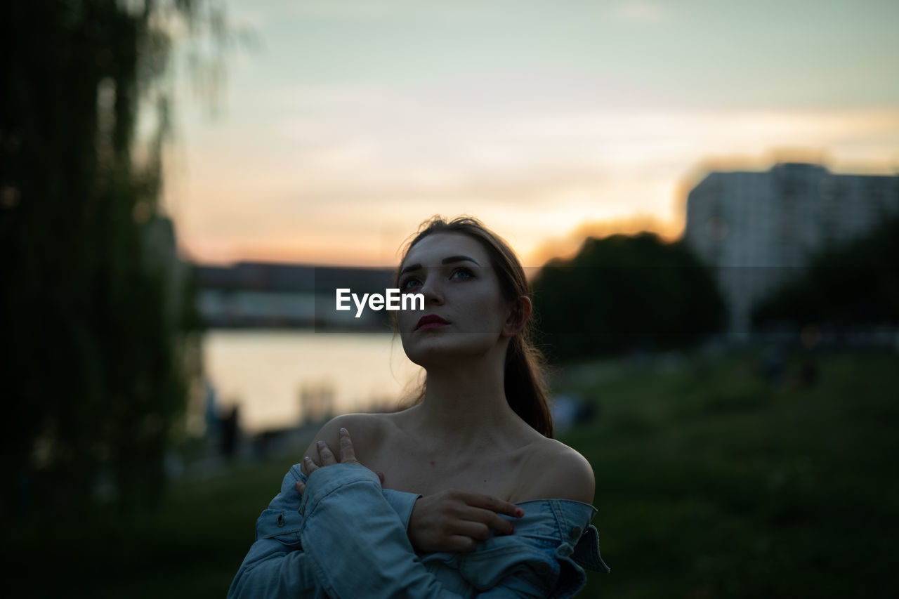 Young woman looking away while standing outdoors during sunset