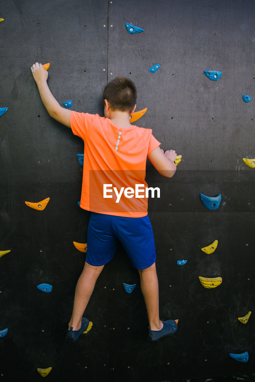 Back view of unrecognizable fearless child balancing on grips and climbing wall while practicing bouldering in gym