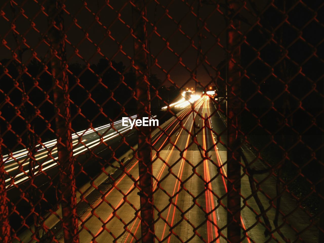 Illuminated vehicle light trails on highway seen from chainlink fence