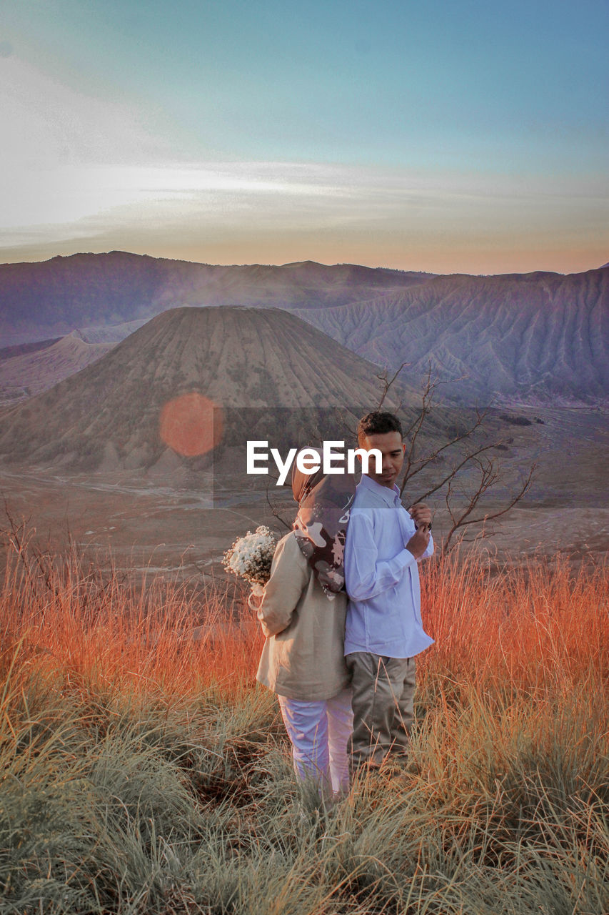 Couple standing on field against sky during sunset