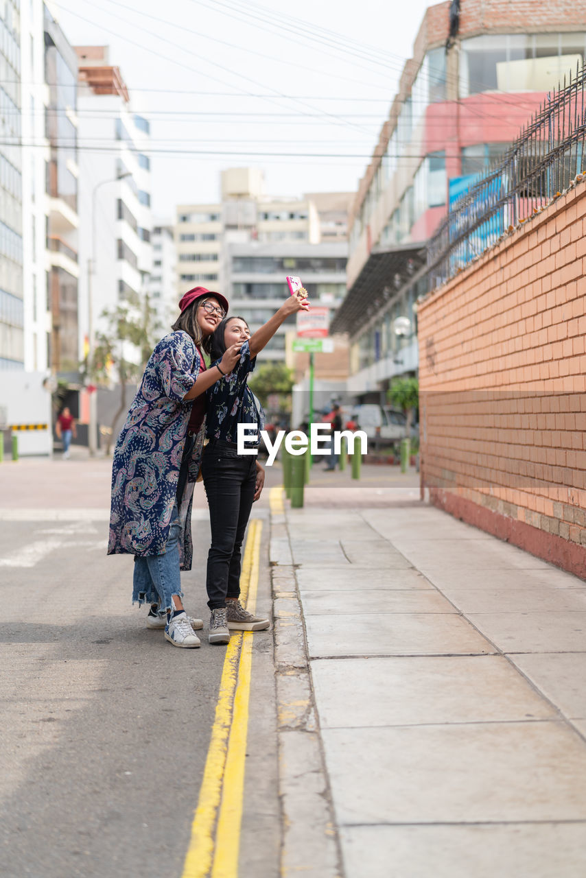 Full body side view of content peruvian female friends taking self portrait on smartphone while standing near road in city