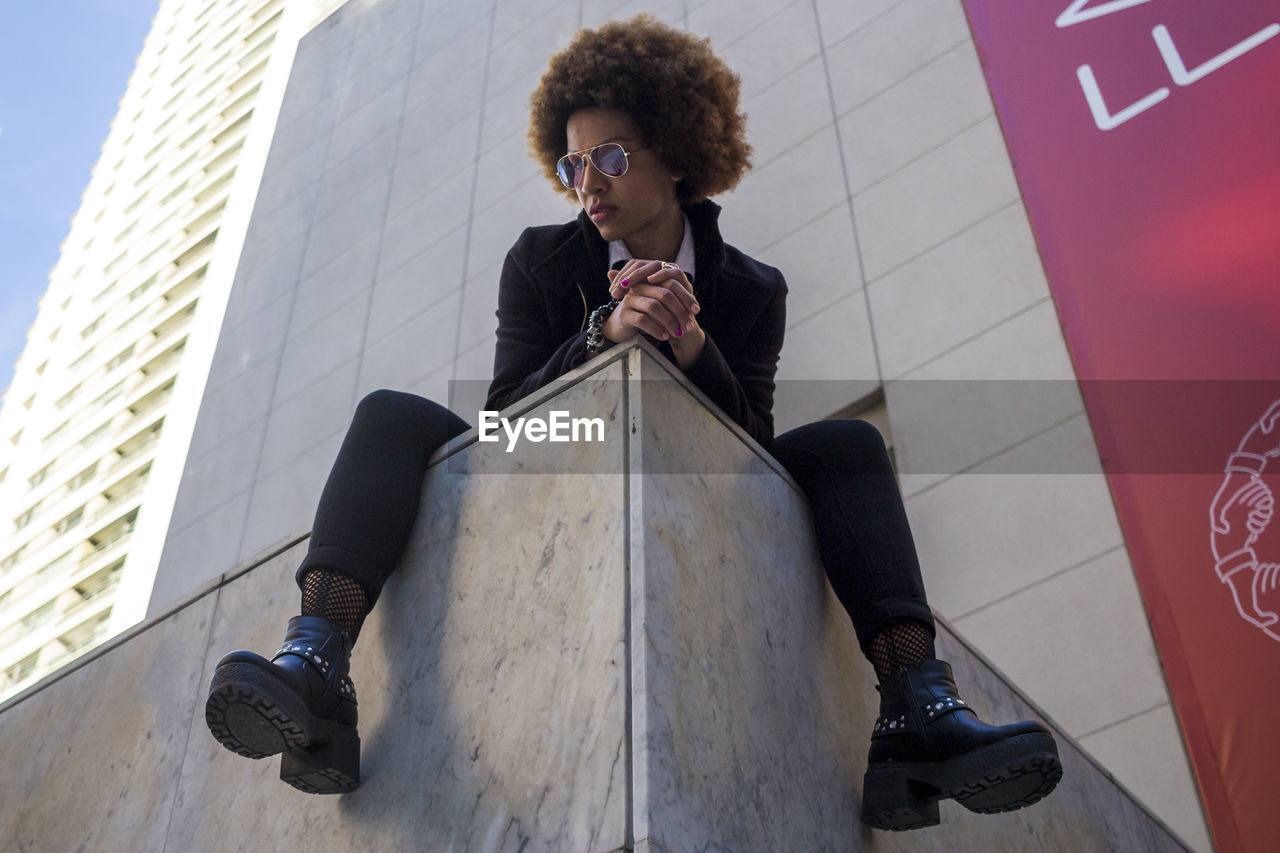 LOW ANGLE VIEW OF YOUNG WOMAN SITTING AGAINST BUILDING