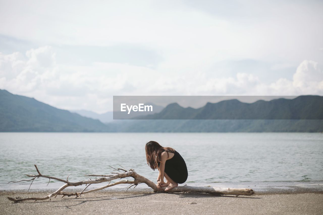 Girl sitting on driftwood log at beach against mountain range