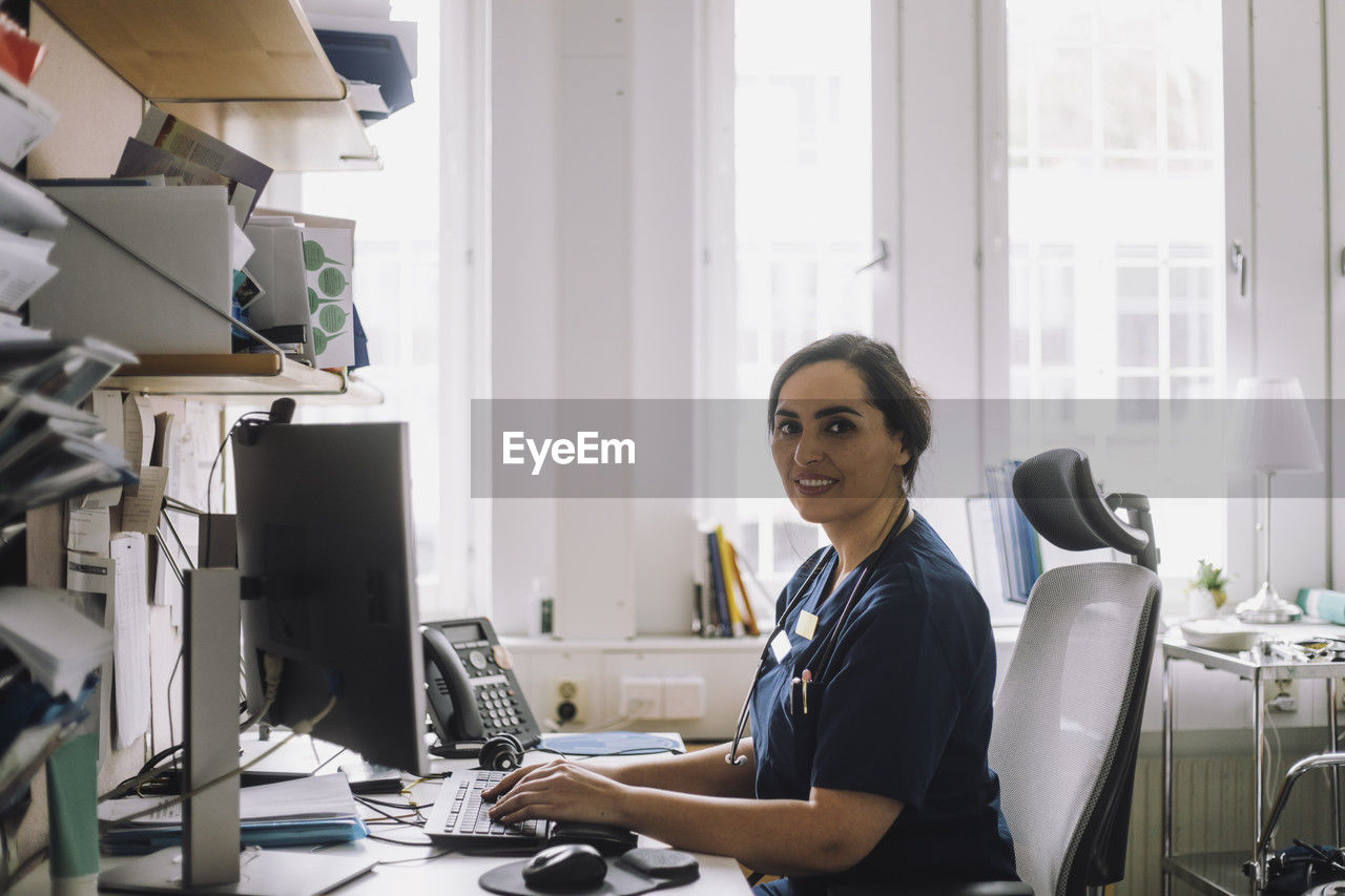 Side view portrait of female nurse sitting at desk with computer in clinic