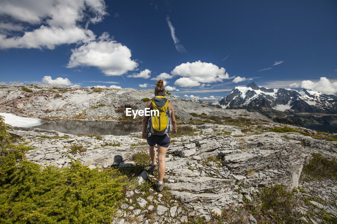 Rear view of female hiker with backpack by pond against mountains and cloudy sky during winter at north cascades national park