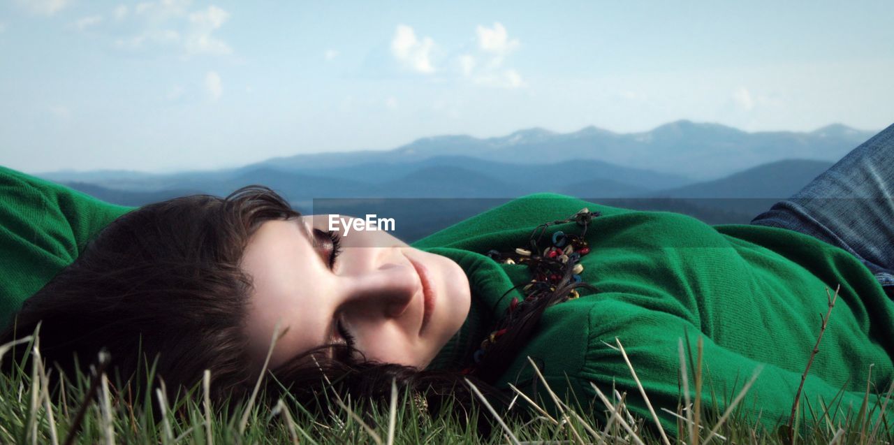 Smiling beautiful woman lying on field by mountains against sky
