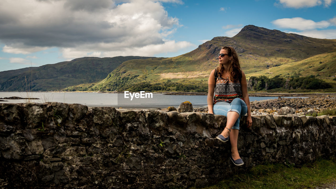 Young woman sitting on retaining wall at lakeshore against mountains