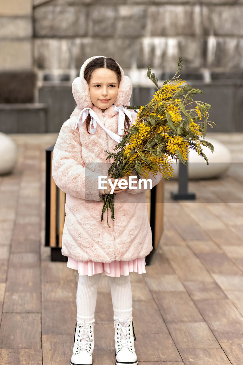 Portrait of a toddler girl in a warm pink coat and fur headphones with a bouquet of mimosa 