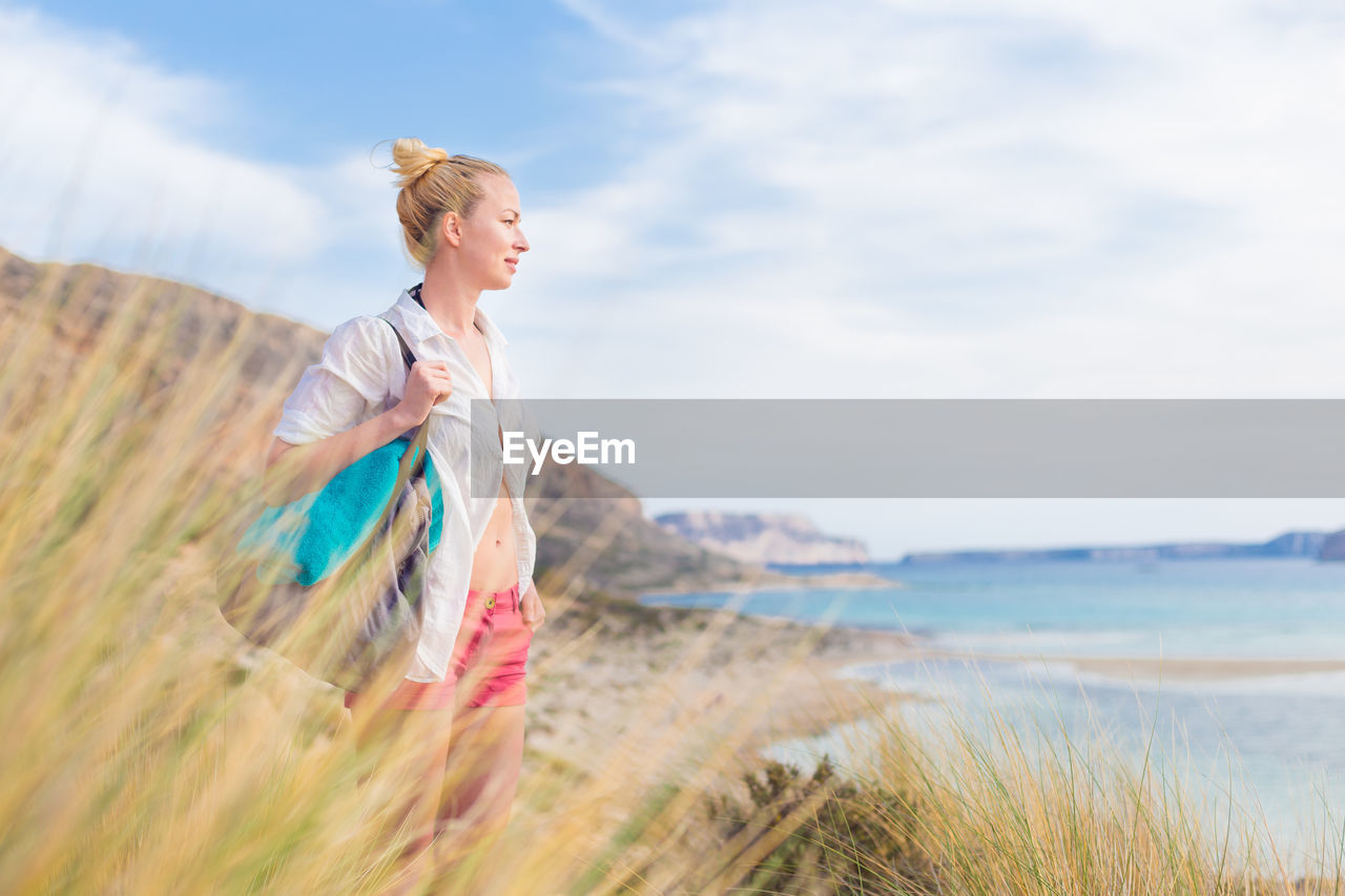 YOUNG WOMAN STANDING ON BEACH AGAINST SKY