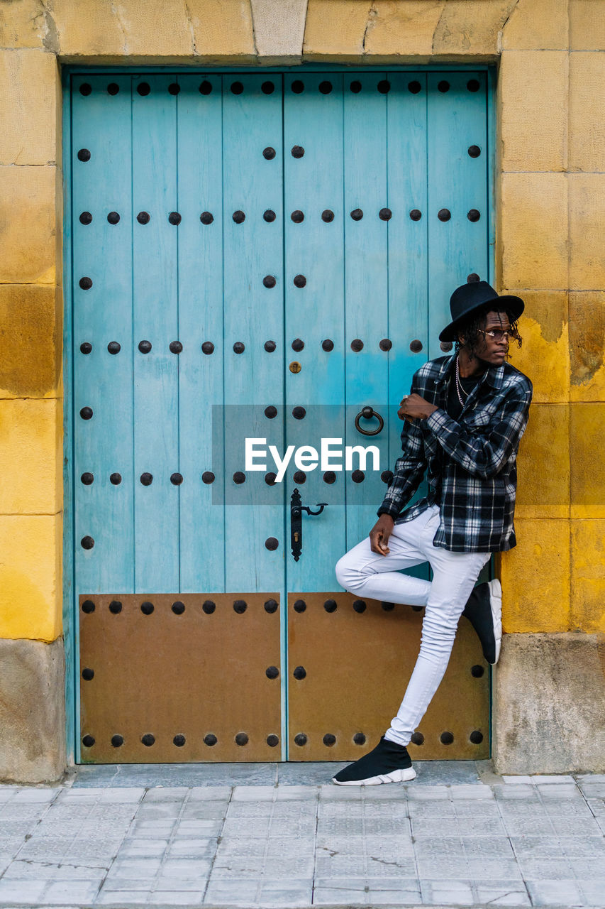 Full body of african american male in trendy outfit and sunglasses standing with crossed legs near doorway