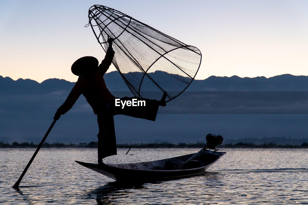 Silhouette man standing on boat in lake against sky during sunset