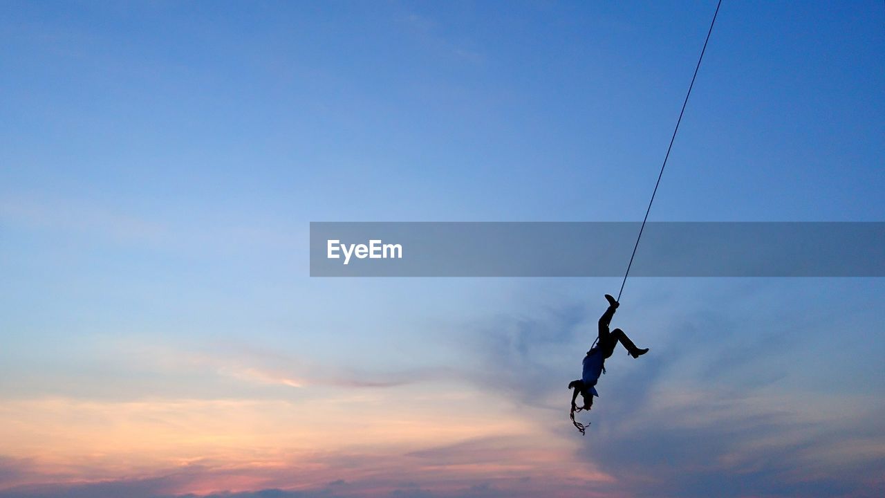 Low angle view of silhouette man hanging on rope against sky