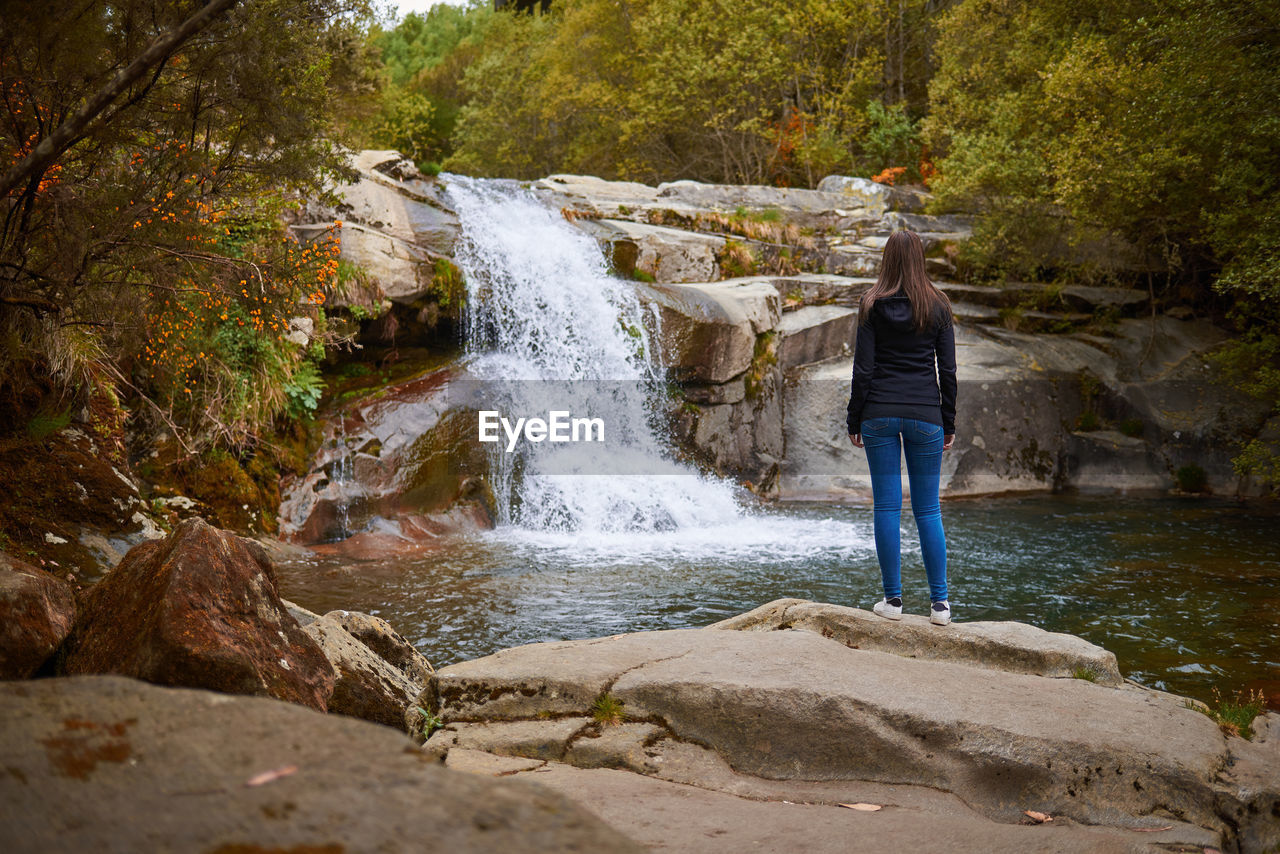 Full length rear view of woman standing on rock against waterfall