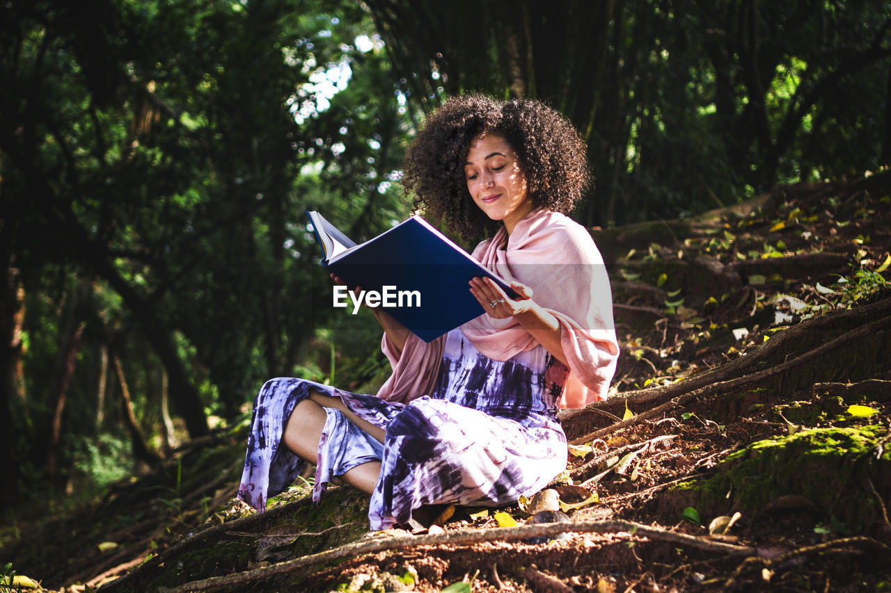 Young woman with a curly hair sitting on a floor with tree roots reading a book and smiling