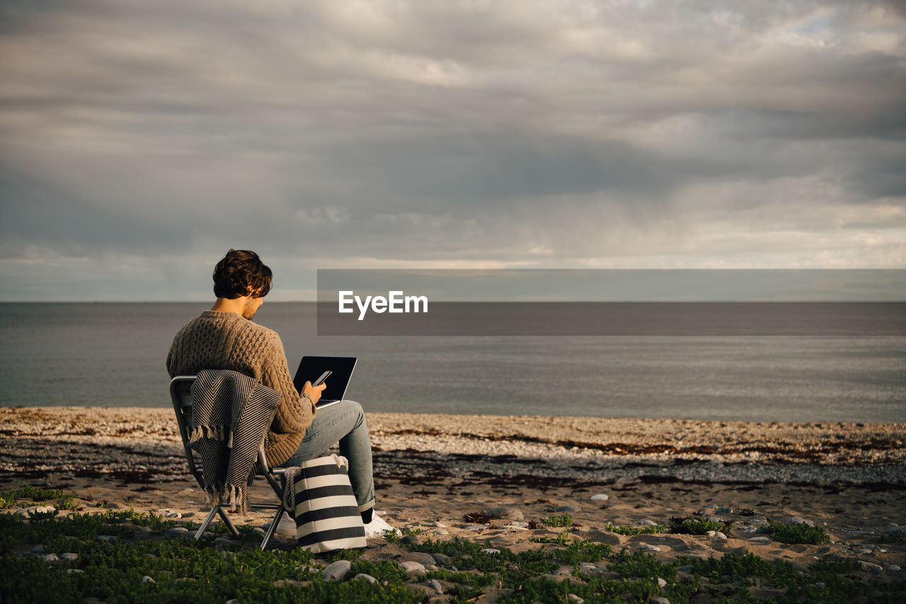 Mid adult man using laptop and mobile phone while sitting on sea shore at beach against sky