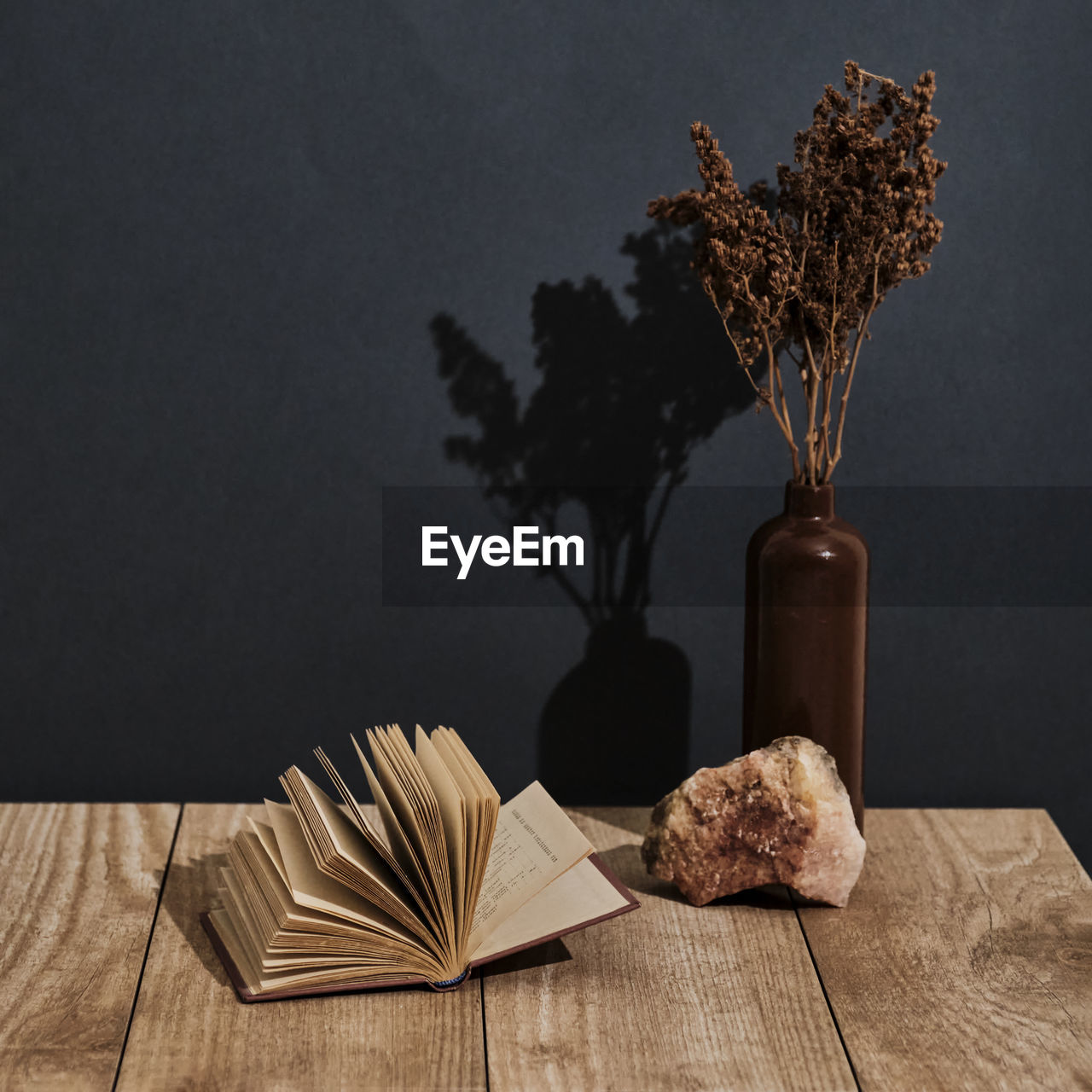 Clay vase with brown dry flowers,natural stone,open old book on wooden table over of black  backdrop