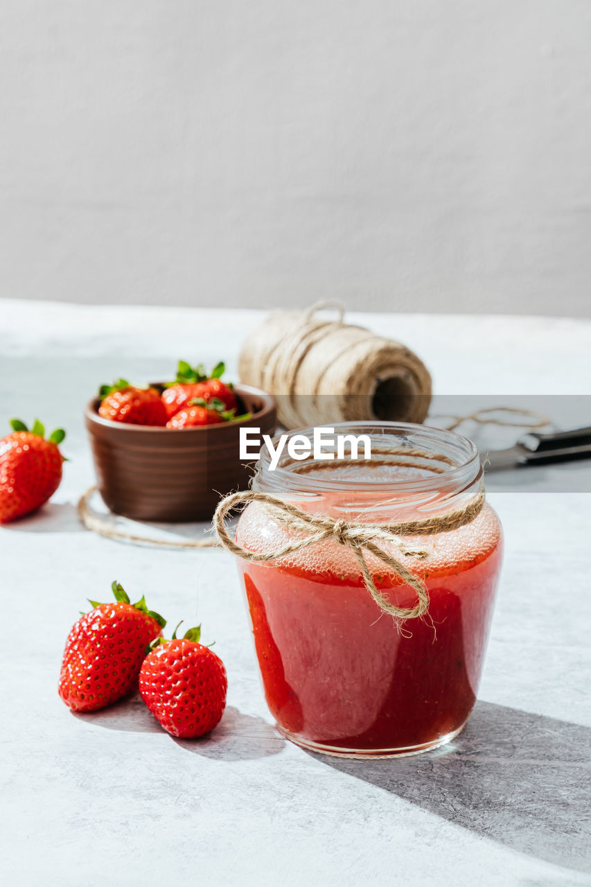 Composition with fresh homemade strawberry juice in glass jar wrapped with twine placed on marble surface with whole berries and knife