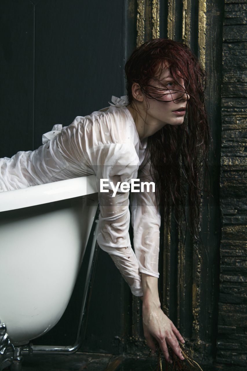 YOUNG WOMAN LOOKING AT CAMERA WHILE STANDING AGAINST WALL