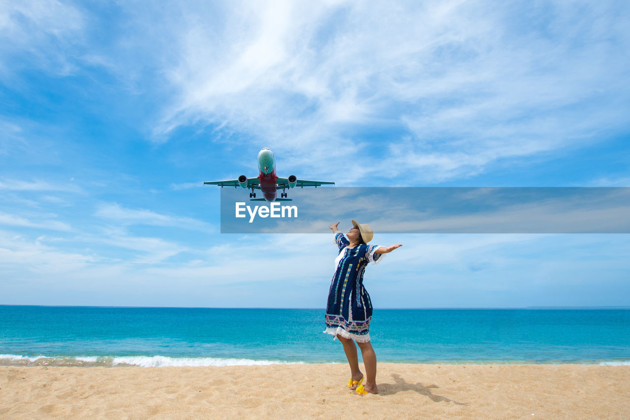 Woman standing below flying airplane at beach against sky