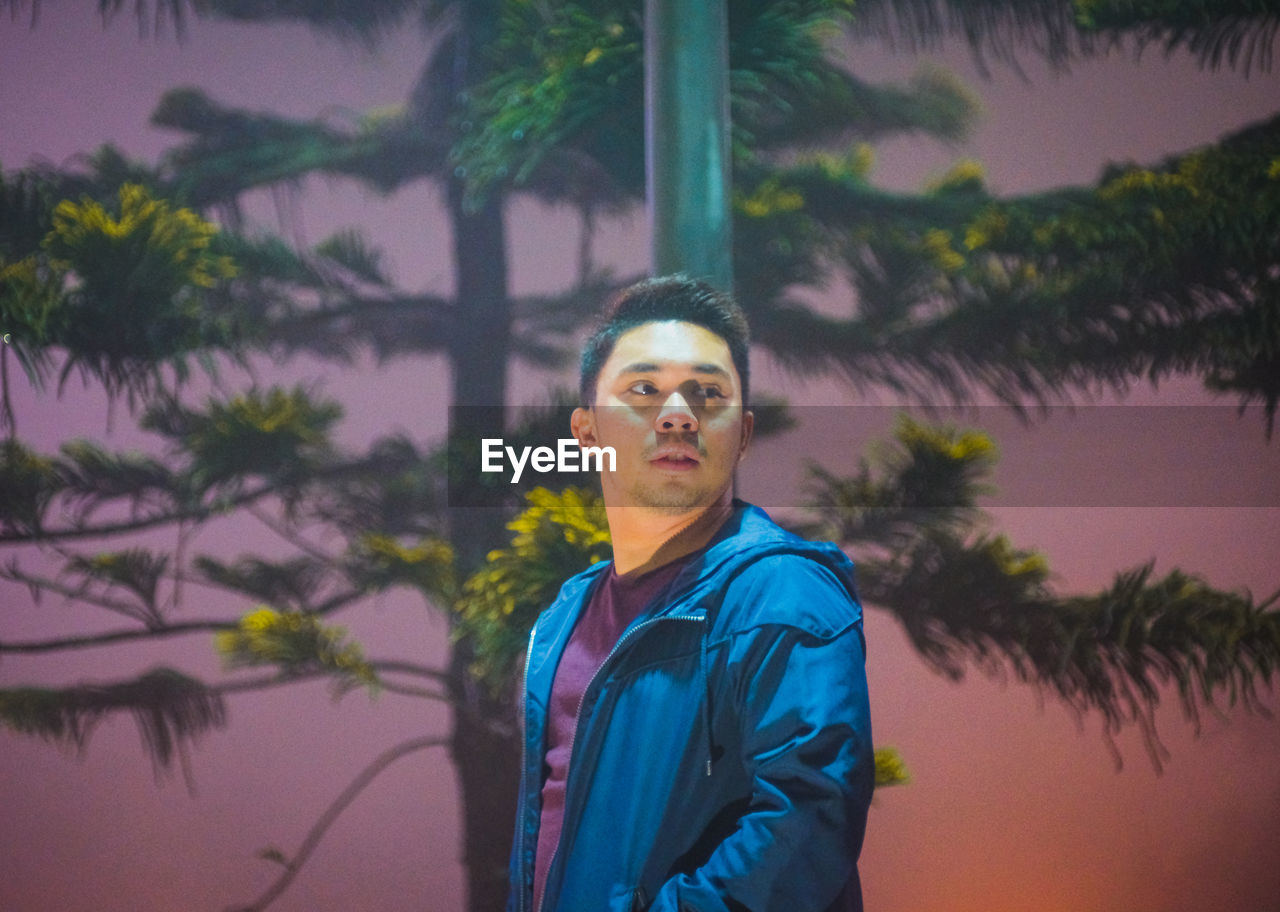 Young man looking away while standing against trees at dusk