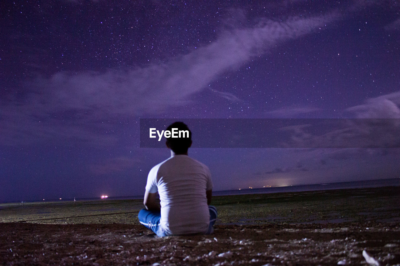 Rear view of man sitting on grassy field against star field at night