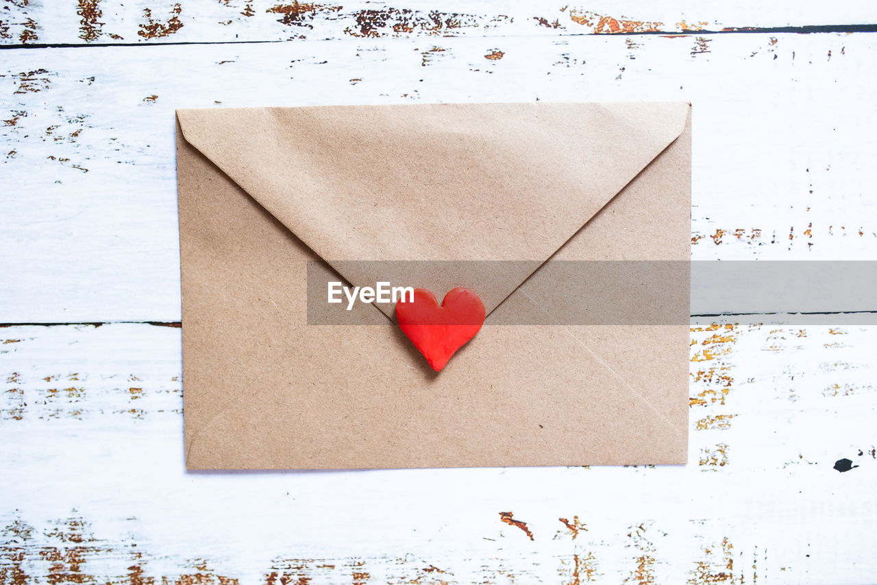 Love letter in a craft envelope with clay red heart on white wood background.