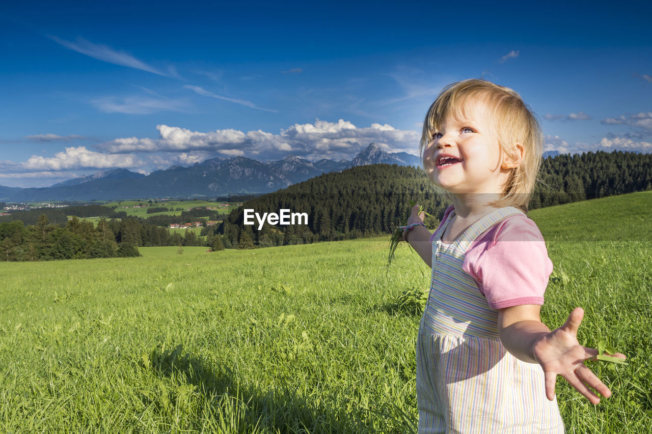 Smiling cute girl gesturing while standing on grass against sky