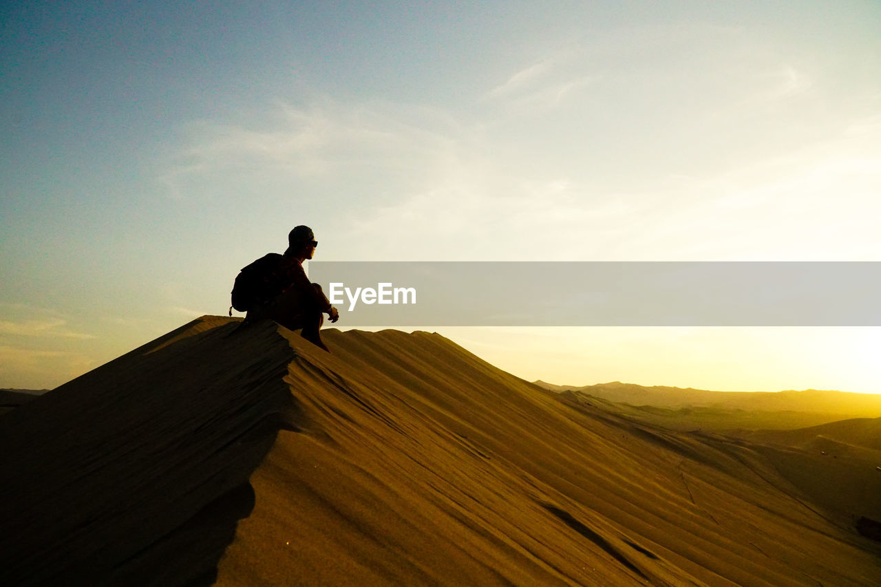 Silhouette man sitting on sand against sky during sunset