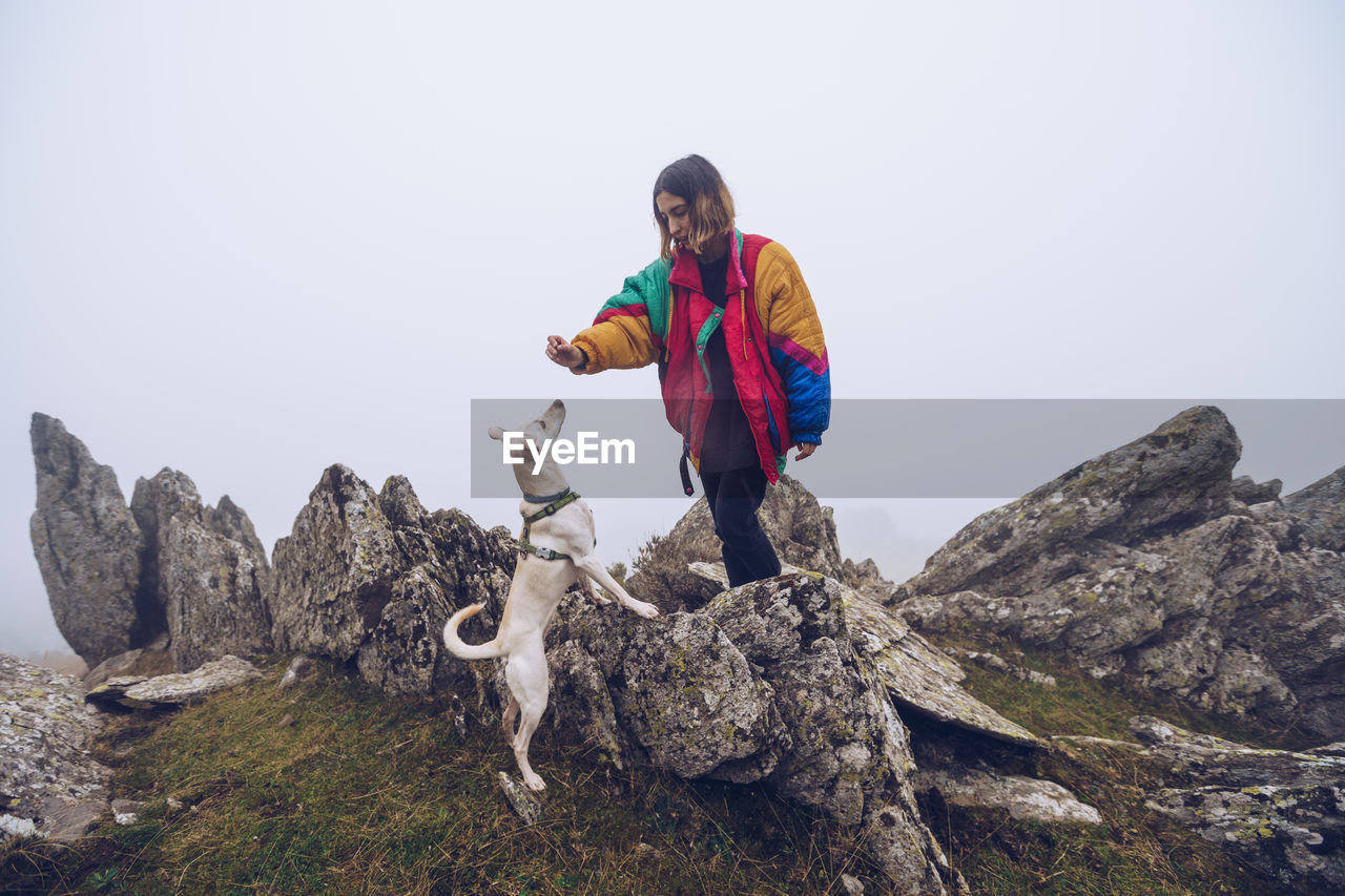 Female owner feeding white dog with treat while standing on rocks in highlands on foggy day