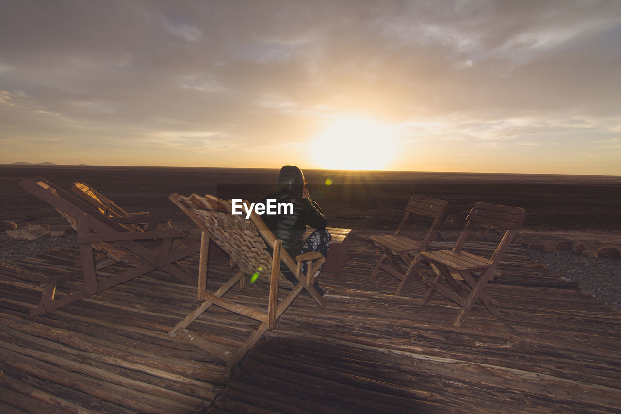 Rear view of person sitting on wooden chair against sky at sunset