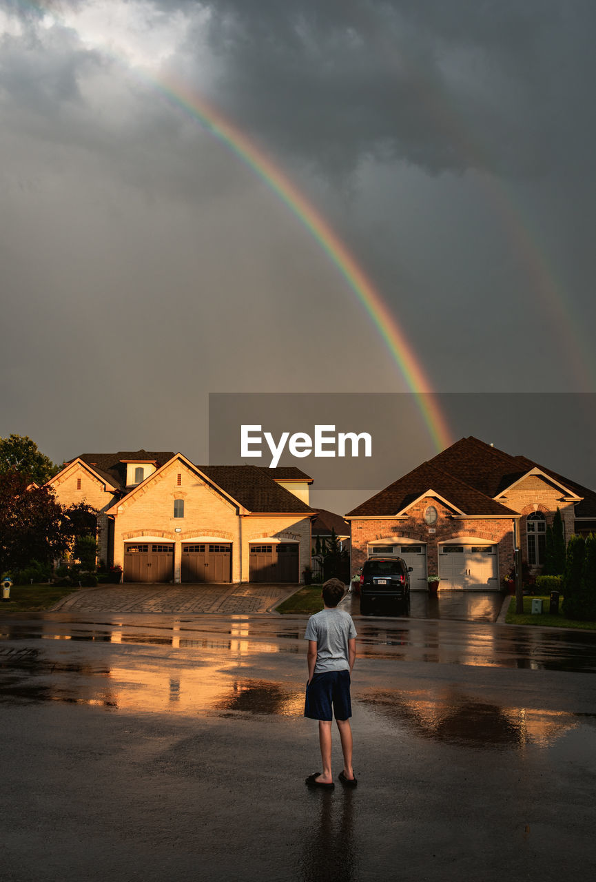 Boy standing in the street looking up at a rainbow in the sky.