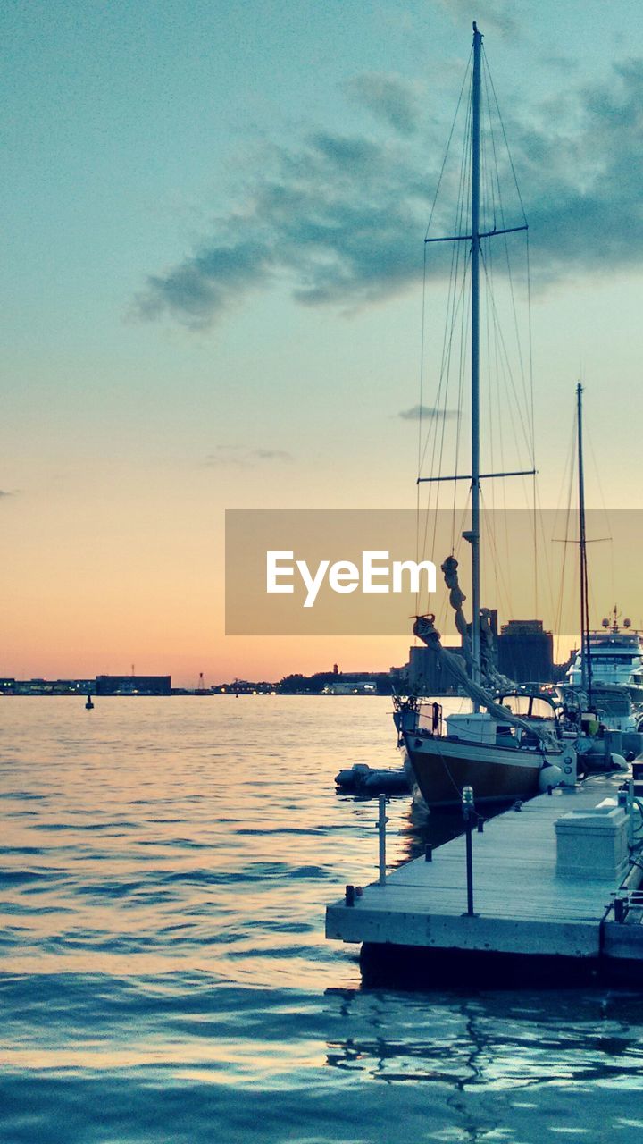 Sailboat moored to pier at harbor against sunset sky