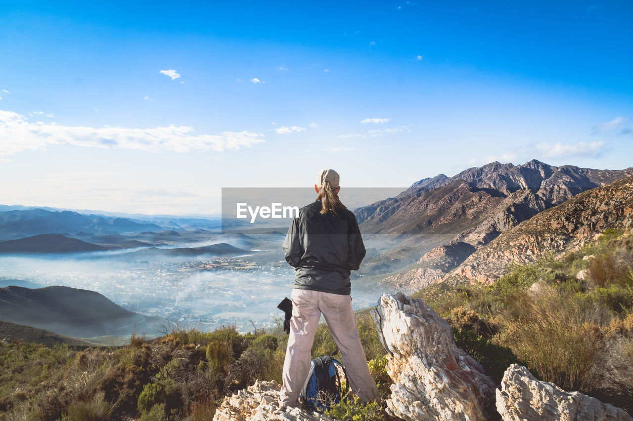 Full length rear view of woman standing on cliff against sky during sunny day