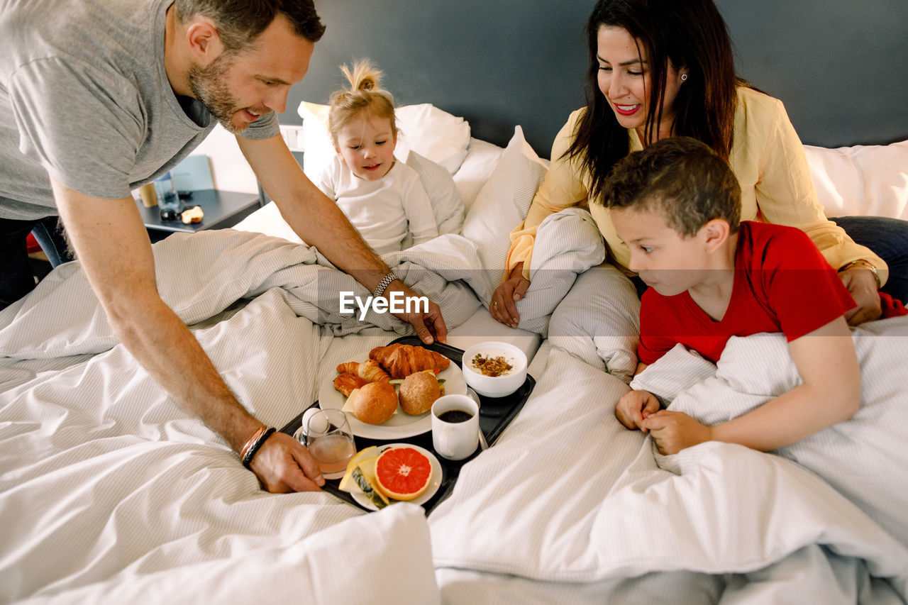 Father serving breakfast for family on bed in hotel room