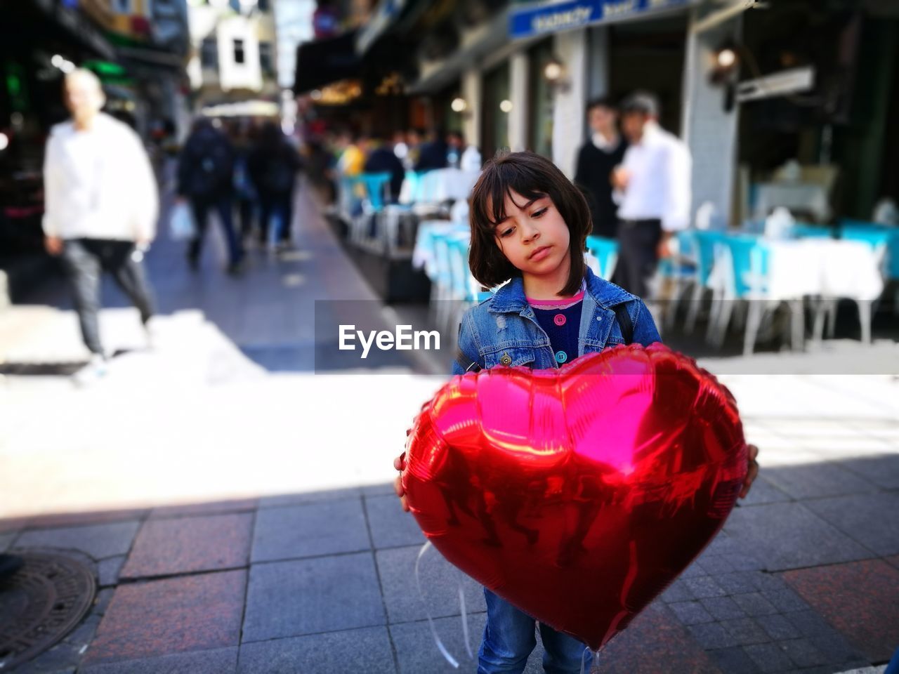 Girl holding heart shape balloon while standing on footpath in city