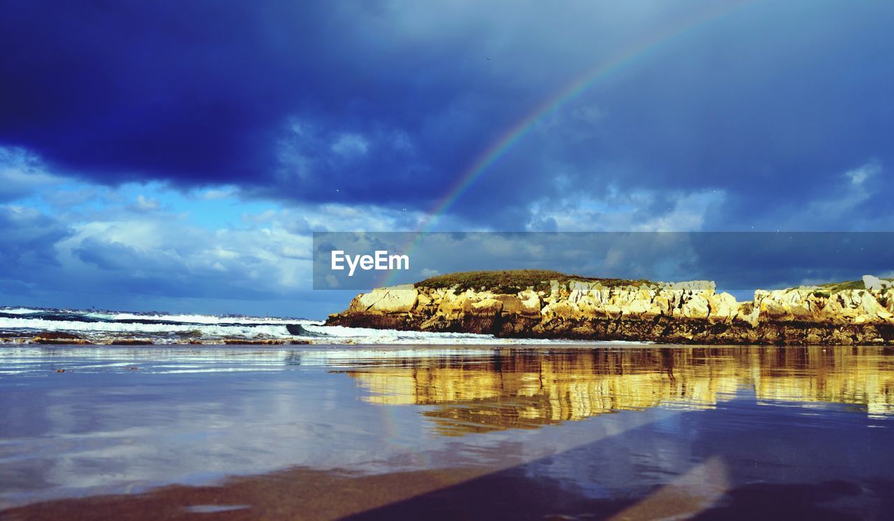 Sea by rocks against rainbow in cloudy sky