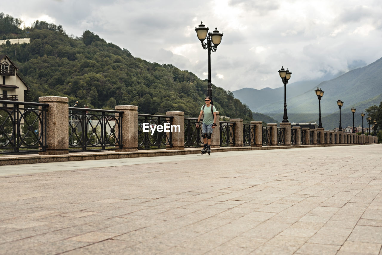 Young man riding on roller skates along embankment against background of forest and mountains 