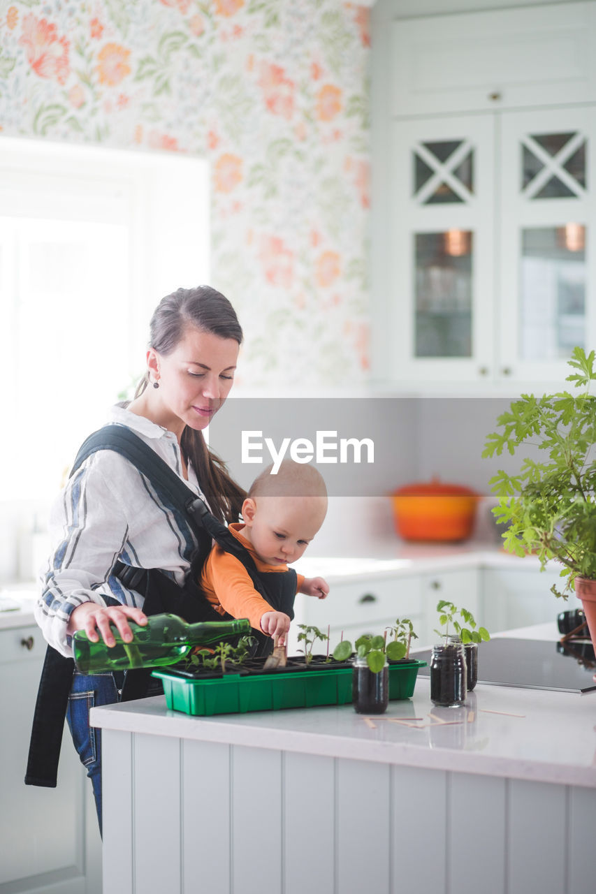 Side view of female blogger watering plants while carrying daughter in kitchen at home