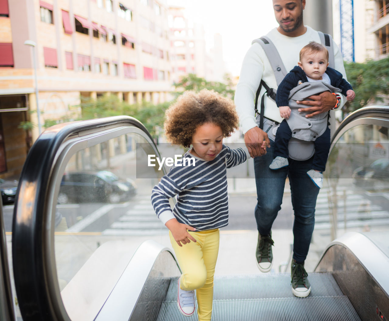 From above of positive young ethnic man carrying infant son and holding hand of cheerful little daughter while standing on escalator in city