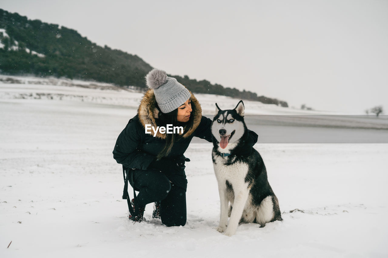 Young ethnic lady wearing outerwear hugging cute husky dog while crouching in snowy woods near green spruces in winter day